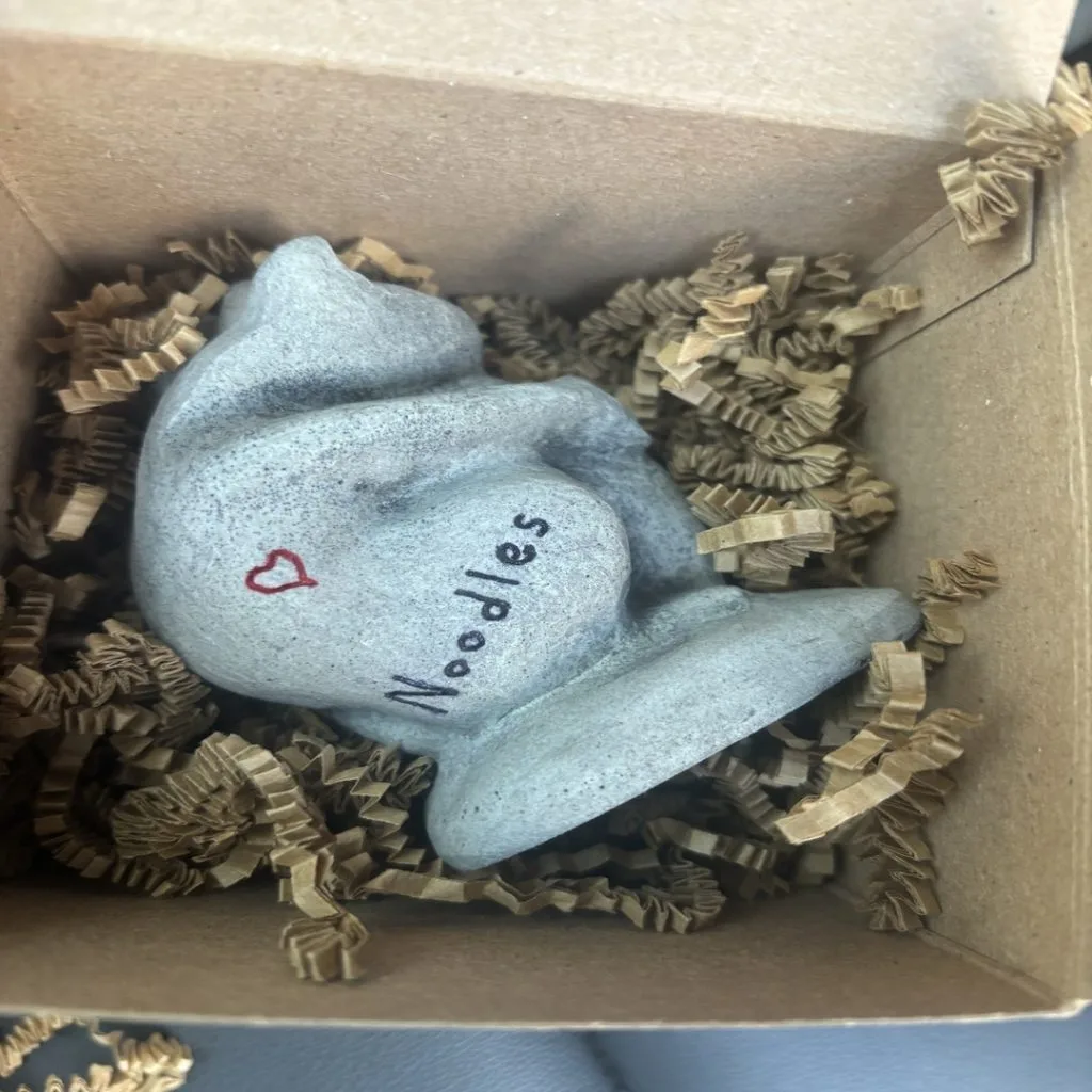 Stone sculpture of a curled-up dog with "Noodles" and a red heart drawn on it, placed in a box with crinkle paper.
